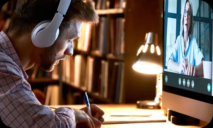 Man sitting at a desk at home, writing notes while in an online tutoring session on his laptop.